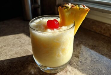 A frosty glass of Iced Pineapple Coconut Mocktail garnished with a maraschino cherry and pineapple wedge.
