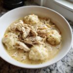 Close-up of a white bowl filled with creamy Instant Pot Chicken and Dumplings, topped with shredded chicken and herbs.
