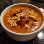 Close-up of a white bowl filled with rich, orange Instant Pot Creamy Chicken Taco Soup, featuring shredded chicken, black beans, and corn.