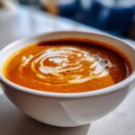 A close-up of a white bowl filled with rich, orange Instant Pot Creamy Tomato Bisque, topped with a swirl of white cream.