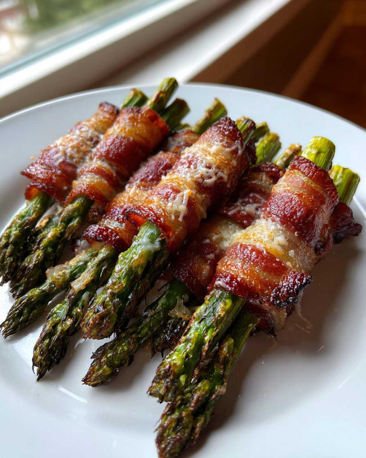 Close-up of perfectly cooked Keto Bacon Wrapped Asparagus Bundles sprinkled with melted cheese on a white plate.