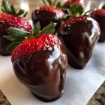 Close-up of a glossy Keto Chocolate Covered Strawberries resting on parchment paper, with more in the background.