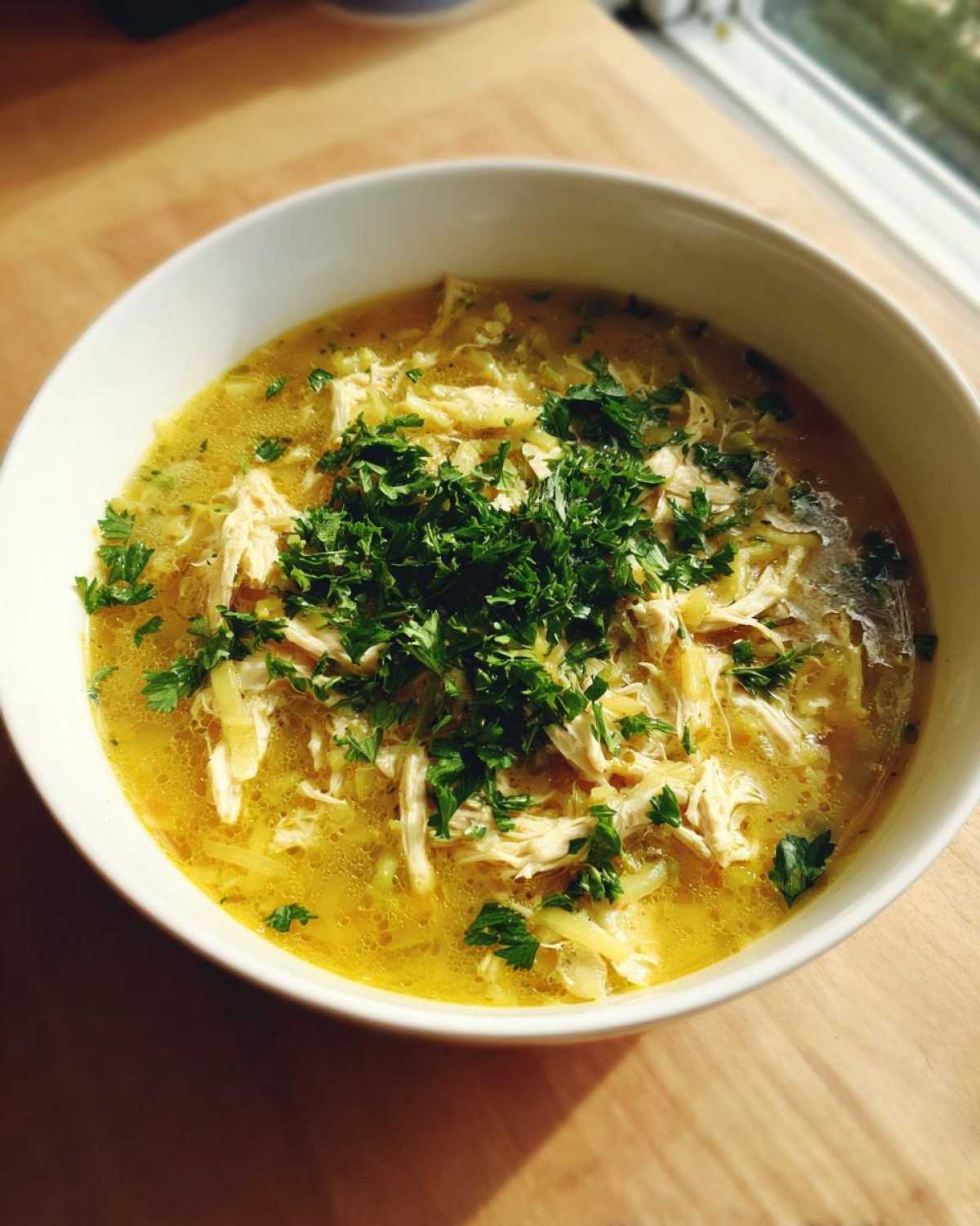 A close-up of a white bowl filled with Keto Creamy Chicken Zoodle Soup, featuring shredded chicken and topped generously with fresh parsley.