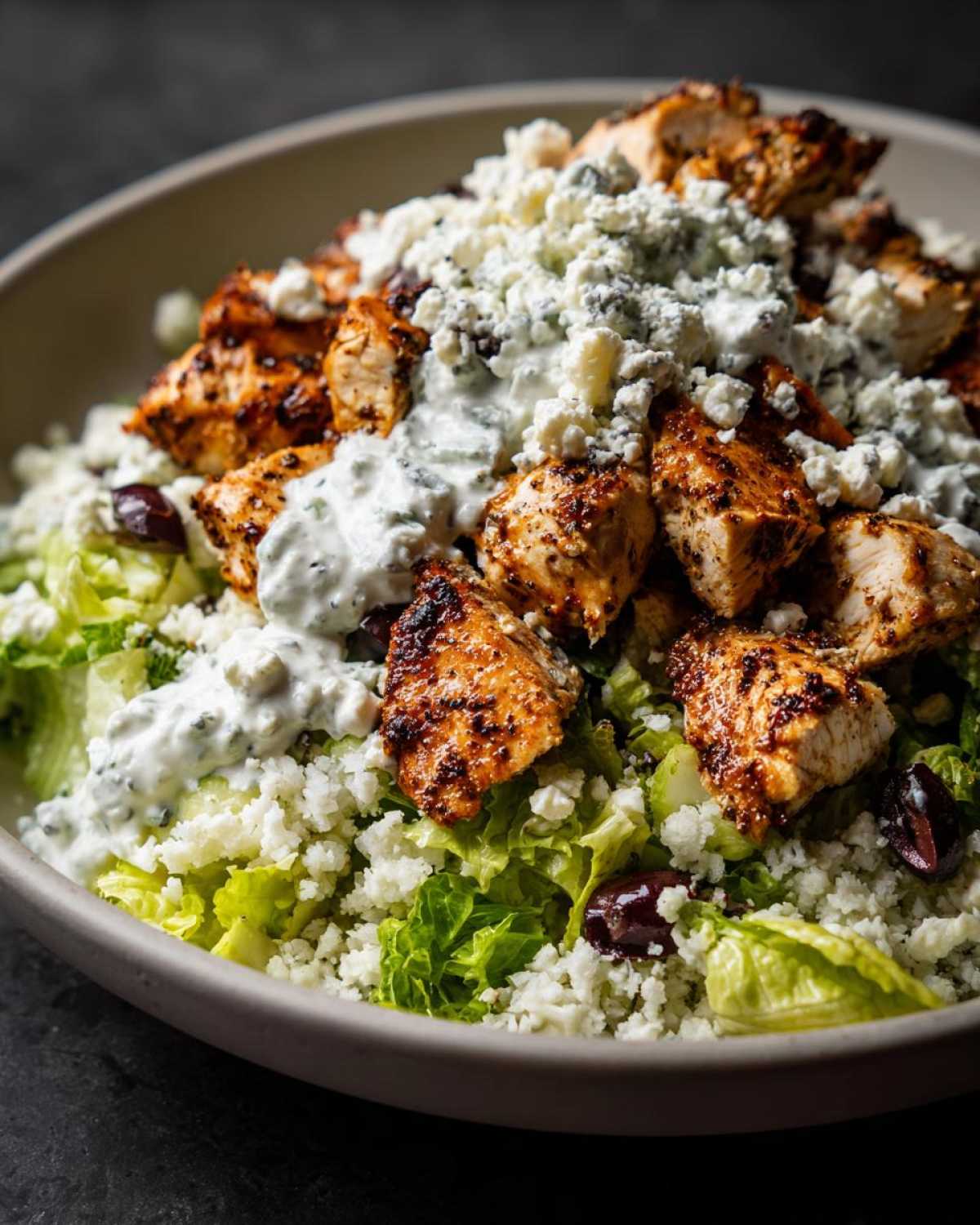 Close-up of a Keto Greek Chicken Bowl featuring seasoned chicken, tzatziki sauce, crumbled feta, and olives over lettuce.