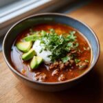 A close-up of a rich, red bowl of Keto Taco Soup with Ground Beef, topped with sour cream, avocado slices, and fresh cilantro.