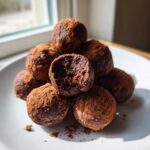 A stack of fudgy No-Bake Brownie Batter Protein Balls dusted heavily with cocoa powder on a white plate.