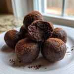 A stack of fudgy No-Bake Brownie Batter Protein Balls dusted with cocoa powder on a white plate.