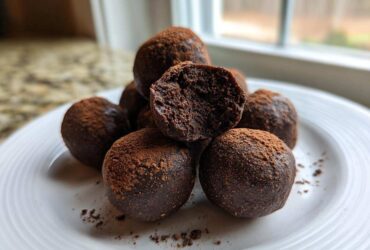 A stack of fudgy No-Bake Brownie Batter Protein Balls dusted with cocoa powder on a white plate.