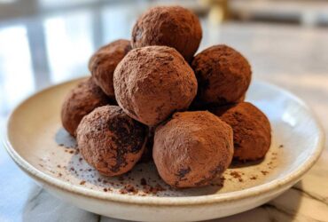 A stack of rich, cocoa-dusted No-Bake Chocolate Chip Cookie Dough Truffles piled on a small, light-colored plate.