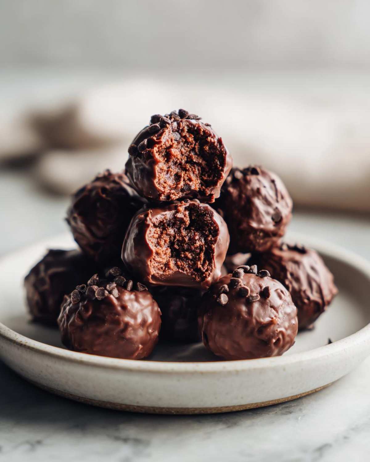 A stack of rich, chocolate-coated No-Bake Chocolate Chip Cookie Dough Truffles, one bitten into, on a light ceramic plate.