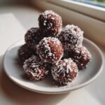 A small stack of rich, dark No-Bake Chocolate Hazelnut Crunch Balls rolled in shredded coconut on a white plate.