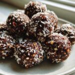 A close-up of several No-Bake Chocolate Hazelnut Crunch Balls rolled in shredded coconut on a light gray plate.