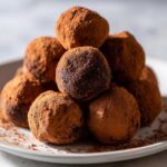 A close-up stack of rich No-Bake Chocolate Orange Truffles heavily dusted with cocoa powder on a white plate.