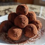 A stack of rich, dark No-Bake Chocolate Orange Truffles generously dusted with cocoa powder on a white plate.