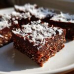 A square piece of No-Bake Coconut Chocolate Date Bars topped with shredded coconut on a white plate.