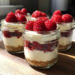 Four individual No-Bake Raspberry White Chocolate Cheesecake Jars layered with graham cracker crust, cream filling, and fresh raspberries.