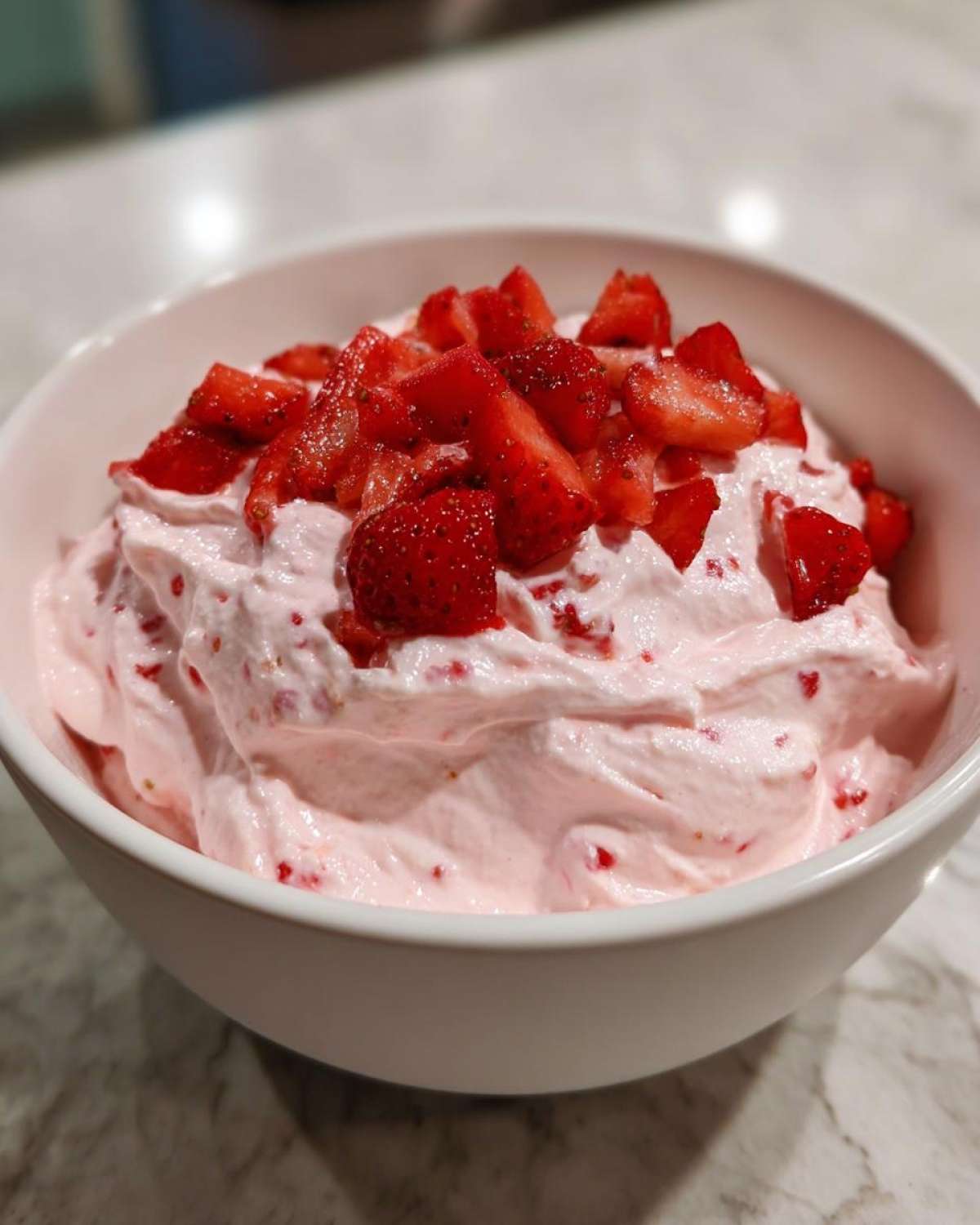 A white bowl filled with pink No-Bake Strawberry Cheesecake Dip, topped generously with fresh, chopped strawberries.