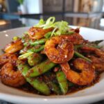 A close-up of glossy, glazed shrimp mixed with bright green snow peas, garnished with scallions, from the One-Pan Honey Garlic Shrimp recipe.