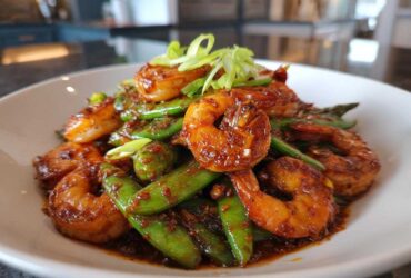 A close-up of glossy, glazed shrimp mixed with bright green snow peas, garnished with scallions, from the One-Pan Honey Garlic Shrimp recipe.