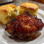 A glazed BBQ chicken thigh served next to two golden cornbread biscuits on a white plate.
