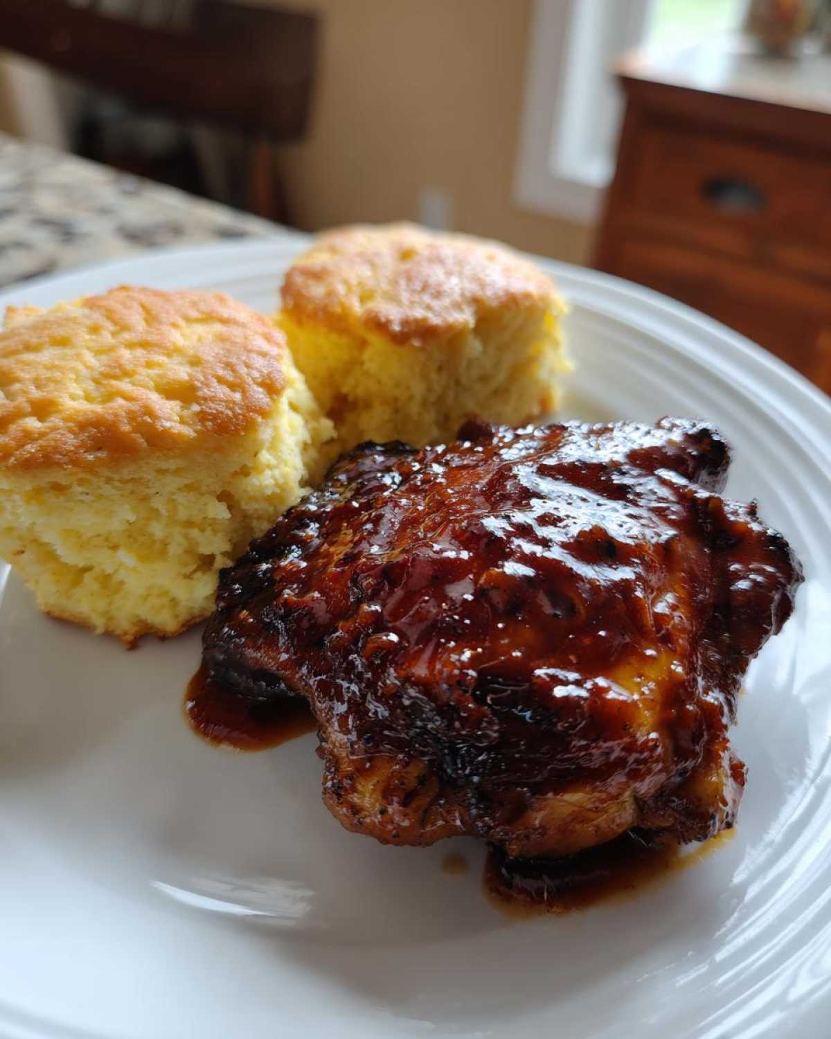 A close-up of a glazed Sheet Pan BBQ Chicken Thighs served next to two golden Cornbread Biscuits on a white plate.