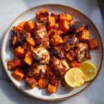 A plate featuring charred Sheet Pan Harissa Chicken with Sweet Potatoes, garnished with lemon slices.