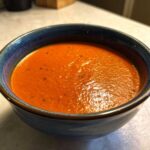 Close-up of a vibrant orange bowl of Vegan Creamy Tomato Coconut Soup, garnished with black pepper specks.