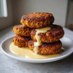 A stack of four golden-brown, crispy Vegan Crispy Chickpea Cutlets drizzled with creamy lemon tahini sauce.