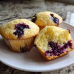 Three moist Vegan Lemon Blueberry Muffins on a white plate, one is cut open showing the soft crumb and burst blueberries.