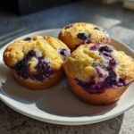 Three freshly baked Vegan Lemon Blueberry Muffins with bright yellow tops and deep purple blueberry bursts, served on a white plate.