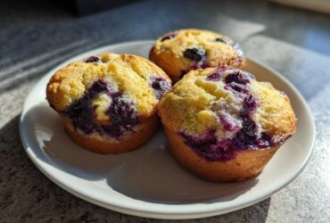 Three freshly baked Vegan Lemon Blueberry Muffins with bright yellow tops and deep purple blueberry bursts, served on a white plate.
