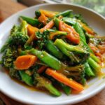 A close-up of vibrant Vegan Spring Vegetable Stir Fry with Ginger featuring bright green snap peas, broccoli florets, and sliced carrots coated in a glossy sauce.