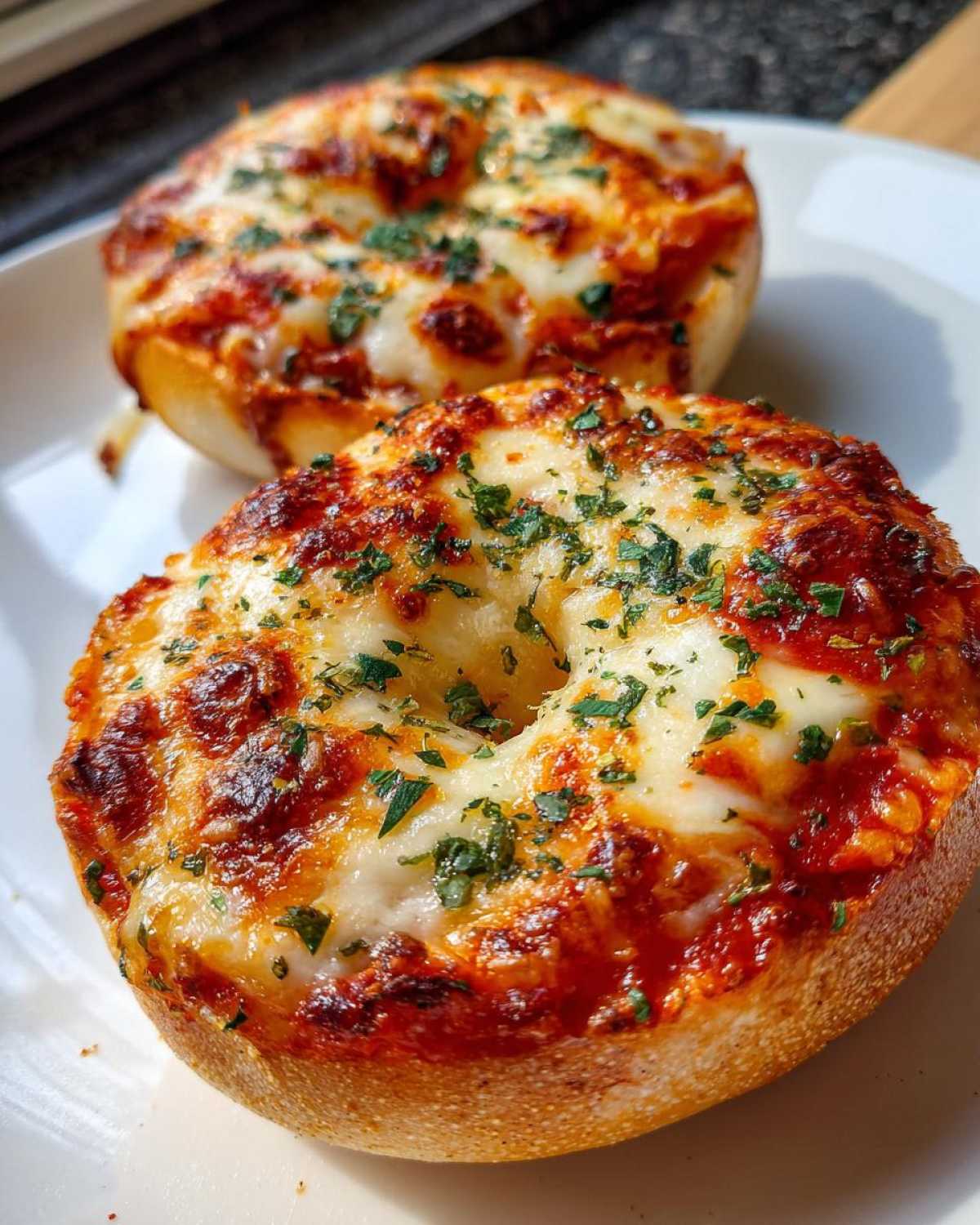 Two perfectly toasted pizza bagels topped with melted, bubbly cheese and sprinkled with green herbs, ready to eat.