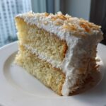A generous slice of a 3-layer coconut cake recipe, featuring moist layers and topped with white frosting and toasted coconut flakes.