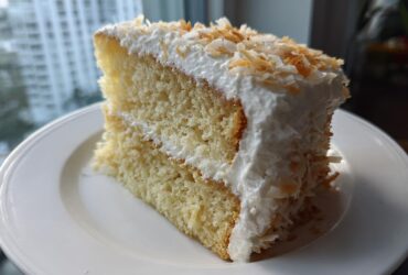 A generous slice of a 3-layer coconut cake recipe, featuring moist layers and topped with white frosting and toasted coconut flakes.