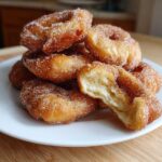 A stack of freshly fried buñuelos coated in cinnamon sugar, with one broken open showing the fluffy interior.