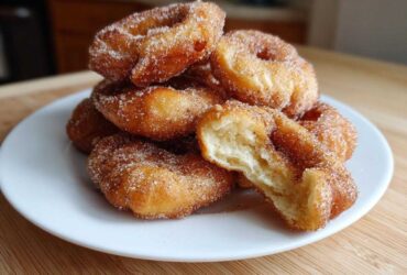 A stack of freshly fried buñuelos coated in cinnamon sugar, with one broken open showing the fluffy interior.