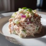 A close-up of a perfectly scooped portion of creamy salmon salad topped with diced red onion and celery.