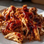 Close-up of shredded bbq chicken generously topped with rich, dark barbecue sauce on a white plate.