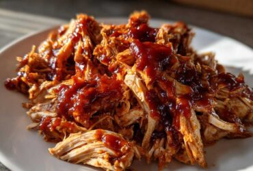 Close-up of shredded bbq chicken generously topped with rich, dark barbecue sauce on a white plate.