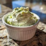 A close-up of creamy, chunky pickle dip topped with dill, served in a small white fluted ramekin.