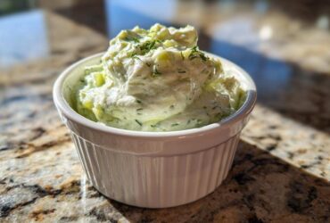 A close-up of creamy, chunky pickle dip topped with dill, served in a small white fluted ramekin.