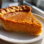 A close-up, sunlit slice of rich orange sweet potato pie on a white plate.