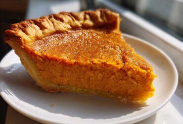 A close-up, sunlit slice of rich orange sweet potato pie on a white plate.