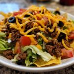 A close-up, appetizing view of a fresh taco salad layered with lettuce, seasoned ground beef, tomatoes, black olives, and shredded cheddar cheese.