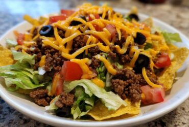 A close-up, appetizing view of a fresh taco salad layered with lettuce, seasoned ground beef, tomatoes, black olives, and shredded cheddar cheese.