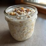A close-up of apple cinnamon overnight oats soaking in milk inside a clear mason jar, topped with apples and cinnamon.