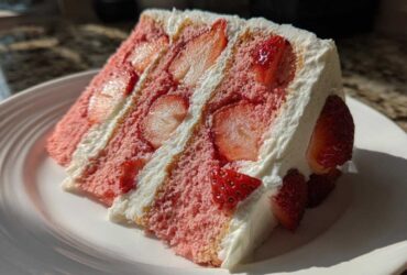 A generous slice of homemade strawberry cake featuring pink layers, fresh sliced strawberries, and thick white whipped cream frosting.