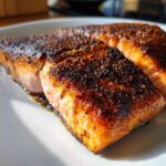 Close-up of a beautifully cooked piece of blackened salmon with a dark, spicy crust on a white plate.