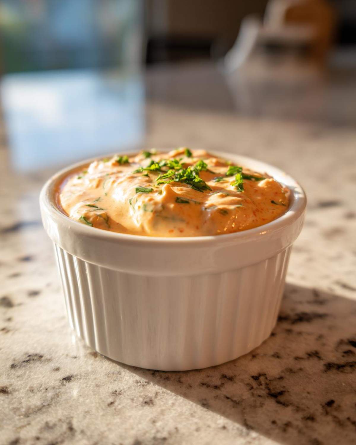 A small white ramekin filled with creamy, orange-hued remoulade sauce, garnished with chopped green herbs.